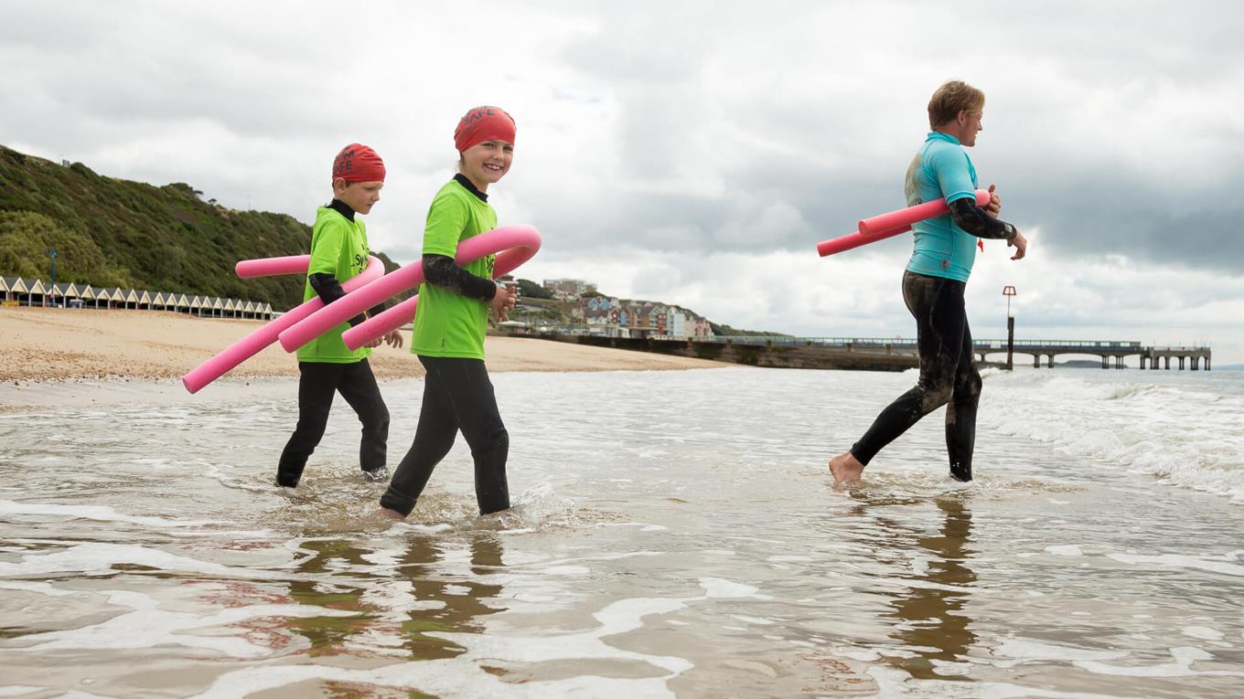 Two children and a swimming instructor in wetsuits walking into the sea