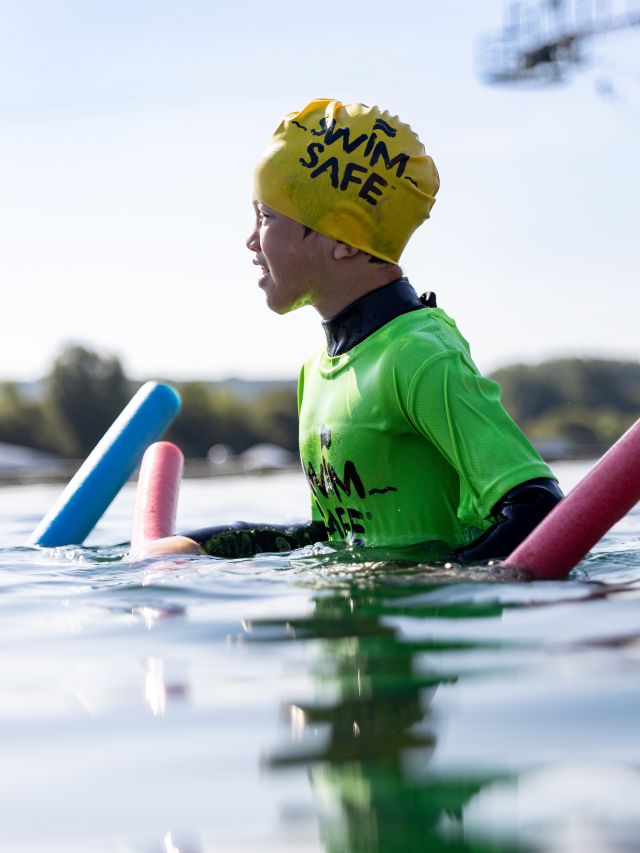 Child wearing a bright green waterproof jacket and yellow patterned hat floats in calm water using pool noodles